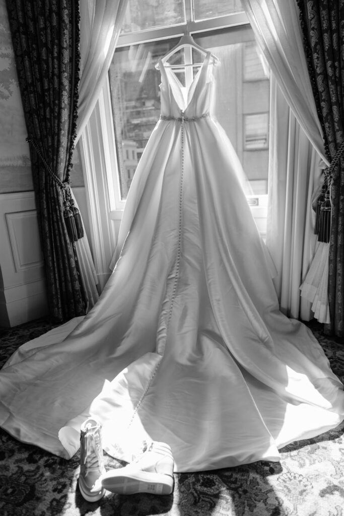 Black and white back lit photo of elegant Pronovias wedding gown in the window of the bridal suite at the University Club NYC photographed by New York City wedding photographer Kat Harris.