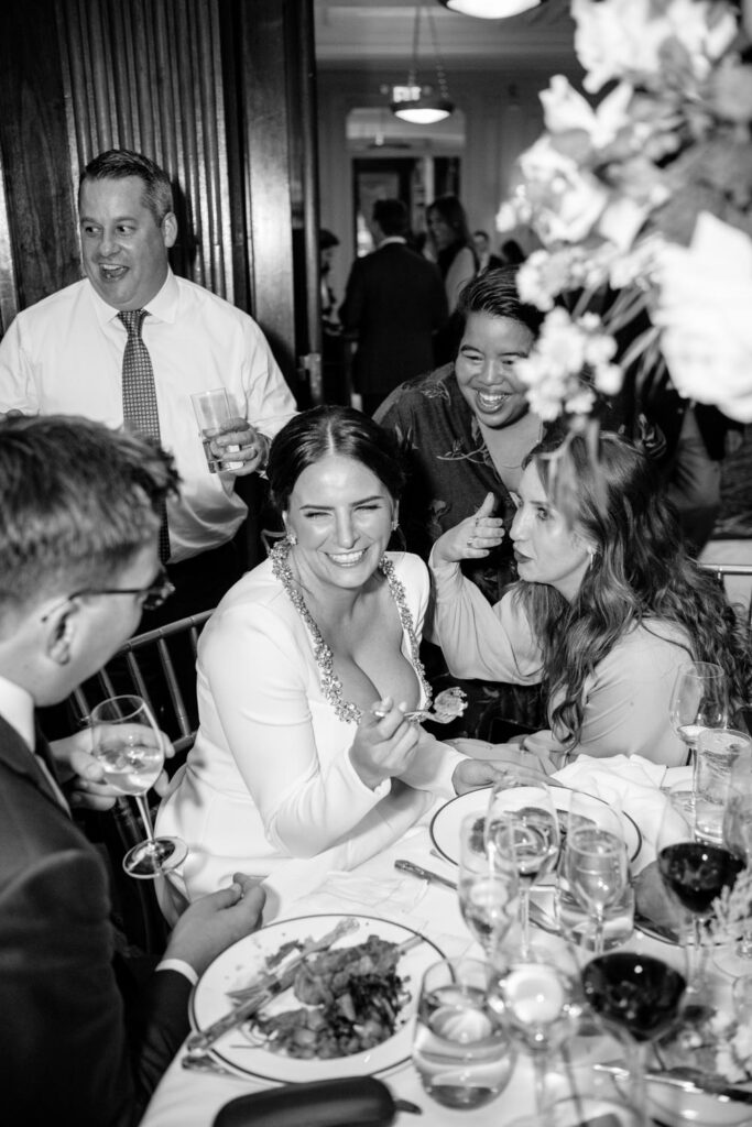 Party photo of bride eating and smiling and laughing while talking with friends during her University Club NY wedding reception photographed by New York City wedding photographer Kat Harris.