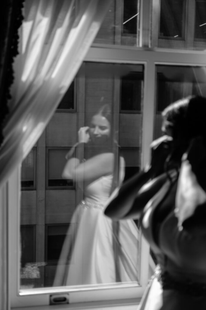 black and white photo of brides reflection in the window while putting on her wedding earrings in the bridal suite before her first look photographed by New York City wedding photographer Kat Harris.