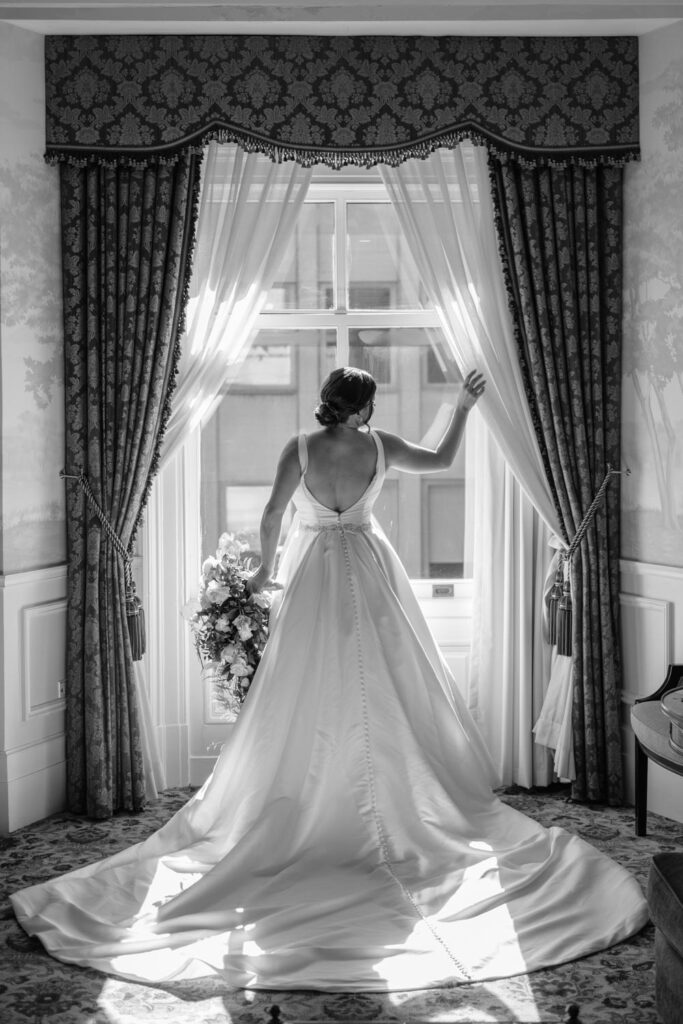 iconic black and white portrait of bride holding her bouquet looking out the window to the busy New York City streets below her as she prepares for her wedding day photographed by New York City wedding photographer Kat Harris.